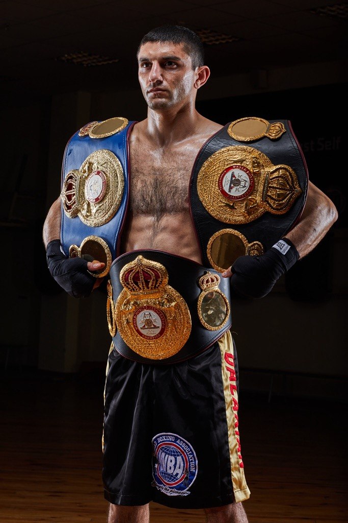 Full length portrait of Artem Dalakian wearing three WBA championship belts and black boxing shorts